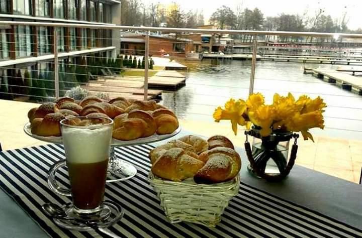 Śniadanie na tarasie z widokiem na jezioro: rogaliki posypane cukrem pudrem na paterze i w koszyku, kawa latte w szklance, żółte narcyzy w wazonie, czarno-biały bieżnik.