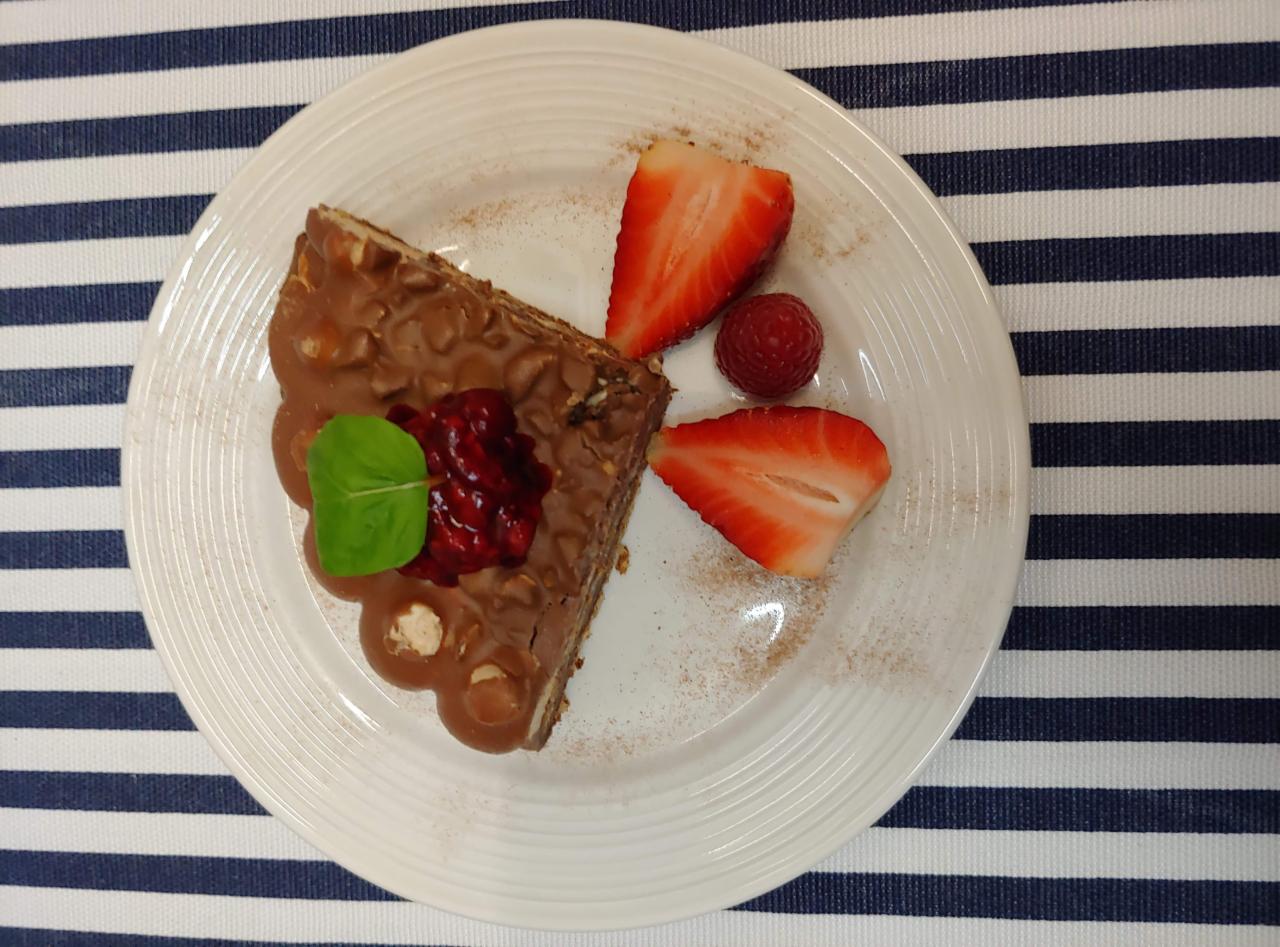 Kawałek tortu orzechowego z polewą czekoladową, udekorowany konfiturą malinową, listkiem mięty, połówkami truskawek i maliną, posypany cynamonem, na białym talerzu z prążkowanym tłem.