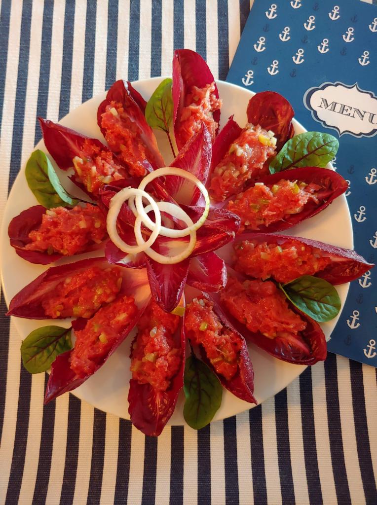 Talerz z cykorią wypełnioną tatarem, udekorowany krążkami cebuli i listkami sałaty, na tle granatowo-białego obrusu w paski i karty menu z motywem kotwic.