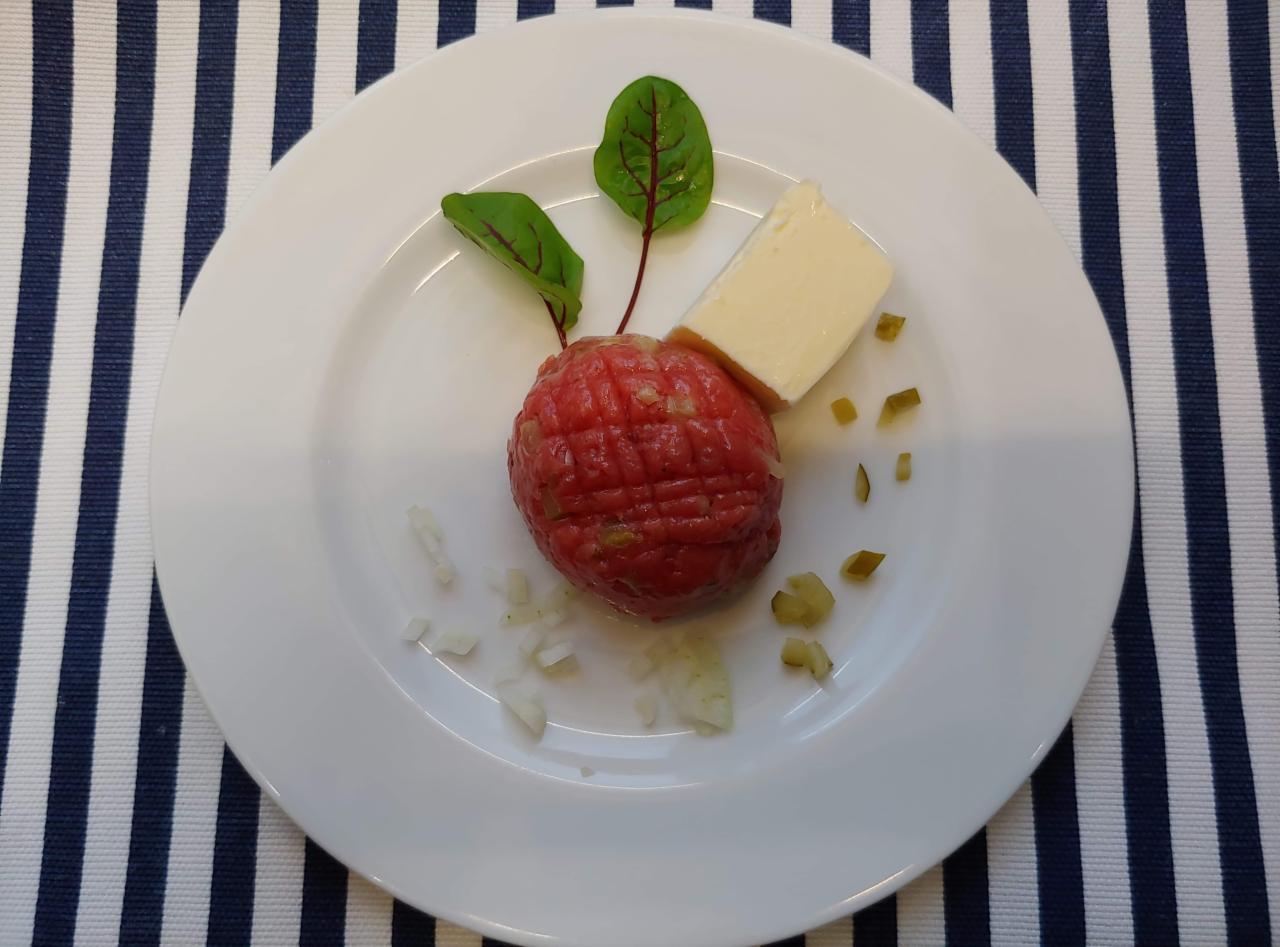 Tatar wołowy uformowany w kulę na białym talerzu, udekorowany dwoma listkami z czerwonymi żyłkami, kostką masła i posiekanymi ogórkami, na tle w biało-granatowe paski.