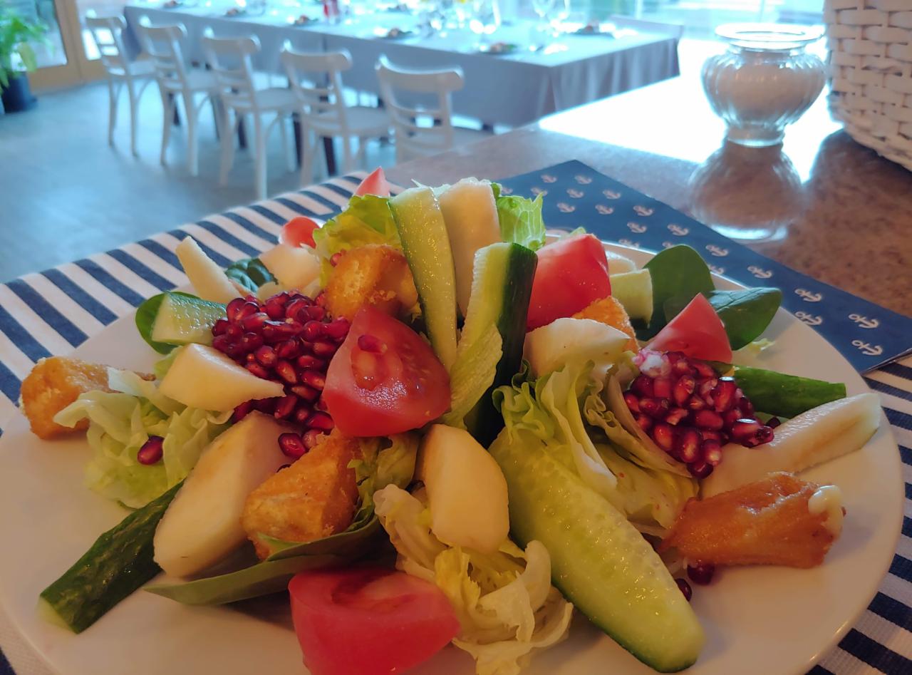 Kolorowa sałatka z granatem, pomidorem, ogórkiem, gruszką i smażonym serem, podana na talerzu z niebiesko-białym obrusem w tle sali z nakrytymi stołami.