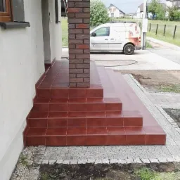 Zewnętrzne schody wejściowe do domu wyłożone terakotą, kolumna obłożona imitacją cegły, widoczny samochód firmy na podjeździe.