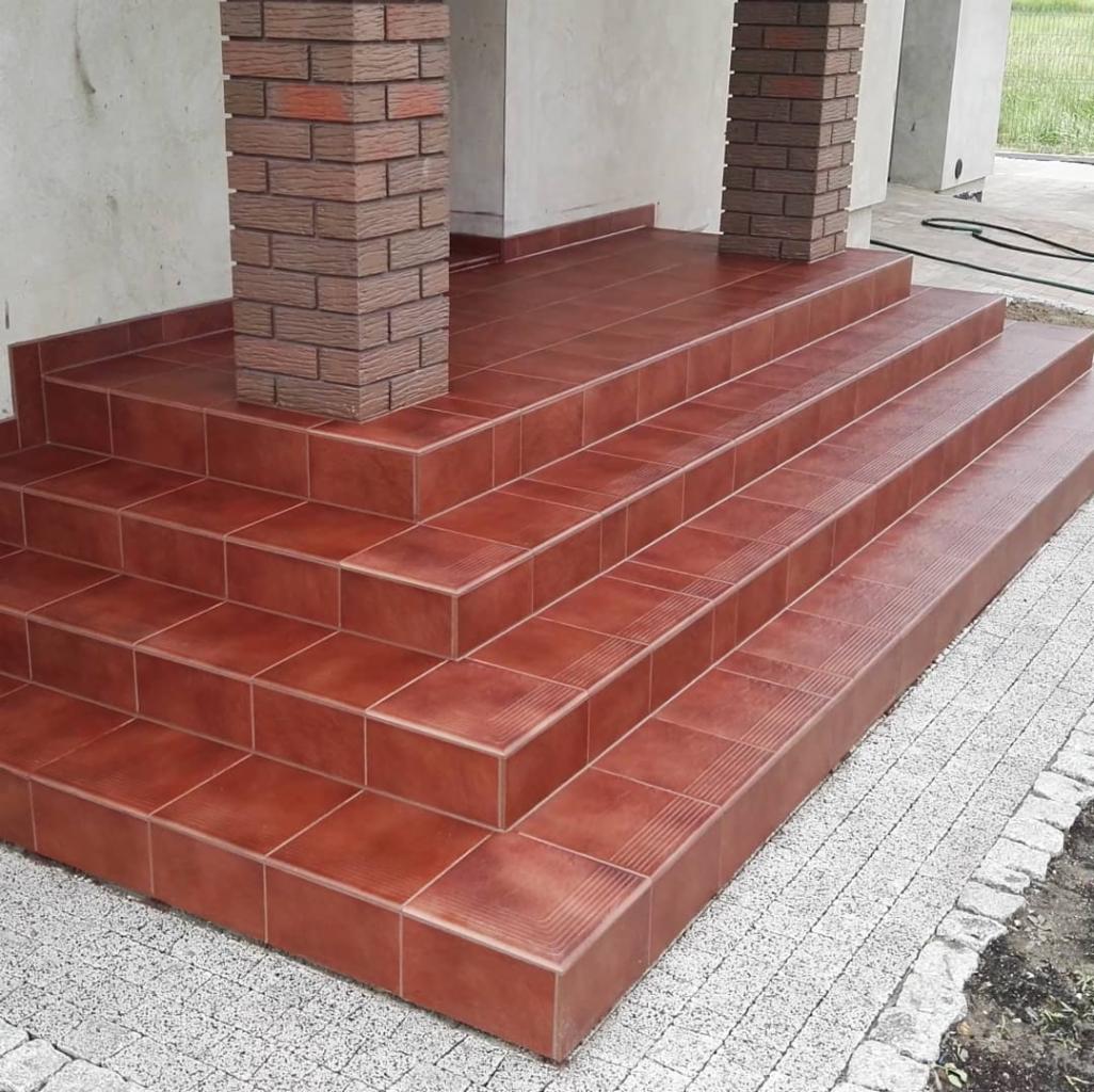Wykończone płytkami schody wejściowe z ceglanymi filarami, widok z dołu ukazujący detale płytek na stopniach i chodniku.