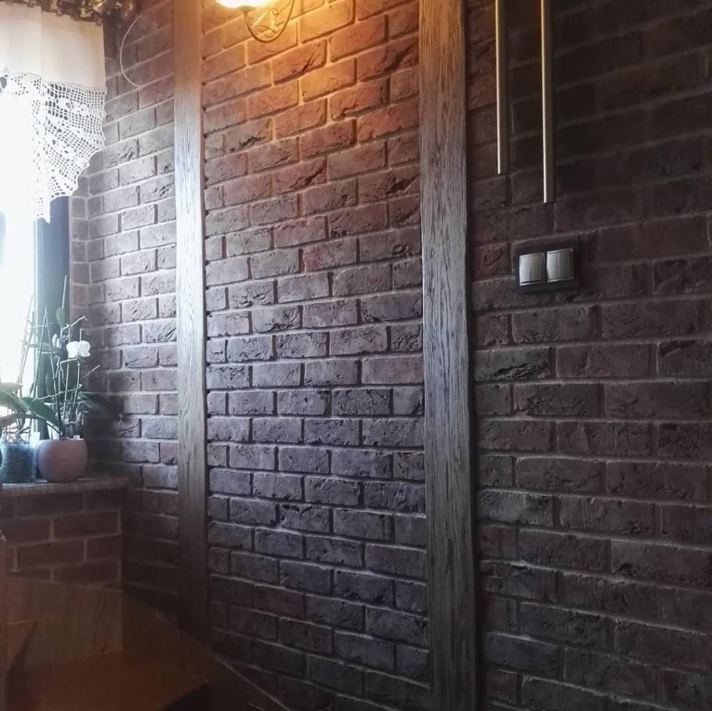 Ściana wykończona dekoracyjną cegłą w odcieniach brązu i szarości, z drewnianą ramą i włącznikiem światła na ścianie, obok okna z koronkową firanką i kwiatami w doniczce.