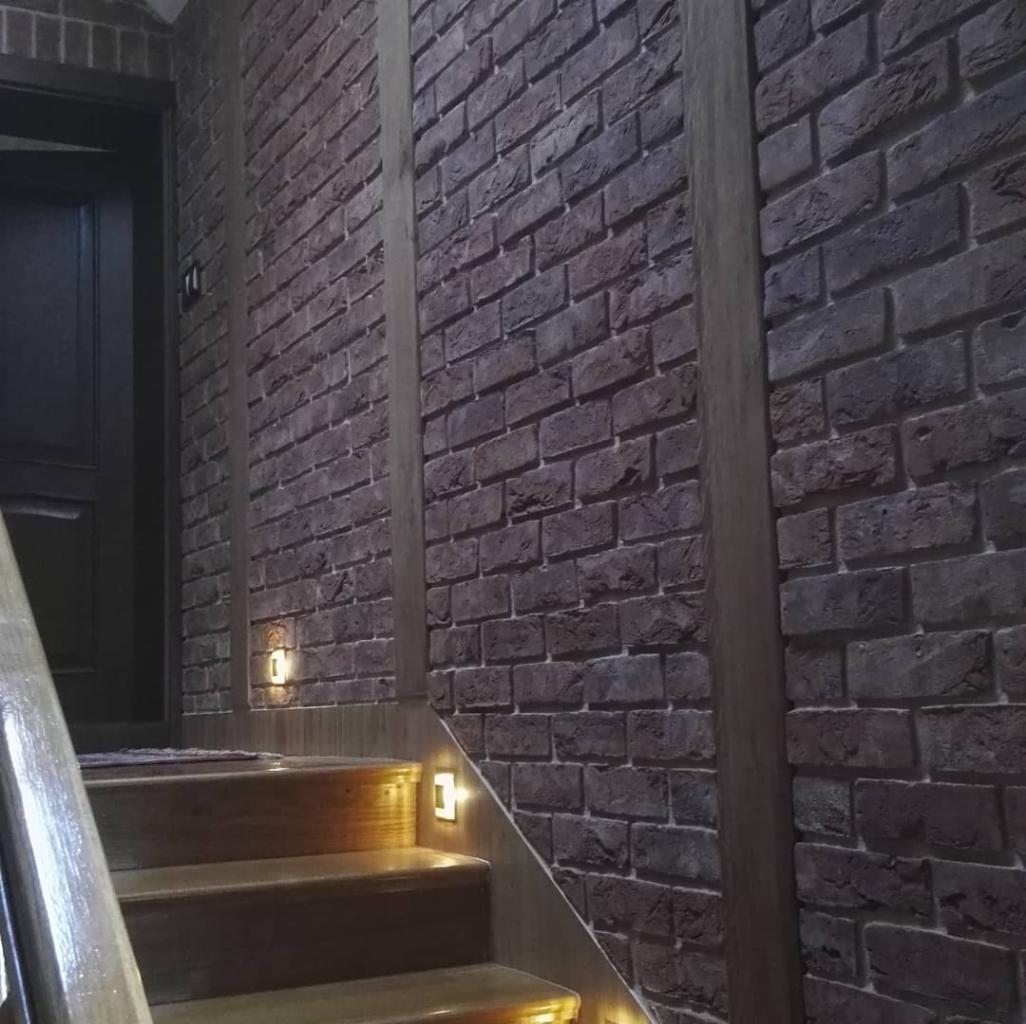 Ciemna ściana z cegły klinkierowej na klatce schodowej, oświetlona małymi, kwadratowymi lampkami LED wbudowanymi w ścianę nad stopniami, z widoczną częścią drewnianej poręczy.