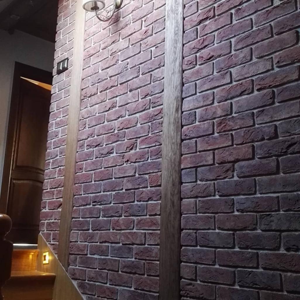 Ściana wewnętrzna wykończona cegłą klinkierową w odcieniach czerwieni i brązu, z fugą w kolorze jasnej szarości, pionowe drewniane listwy dekoracyjne, fragment schodów drewnianych z oświetleniem...