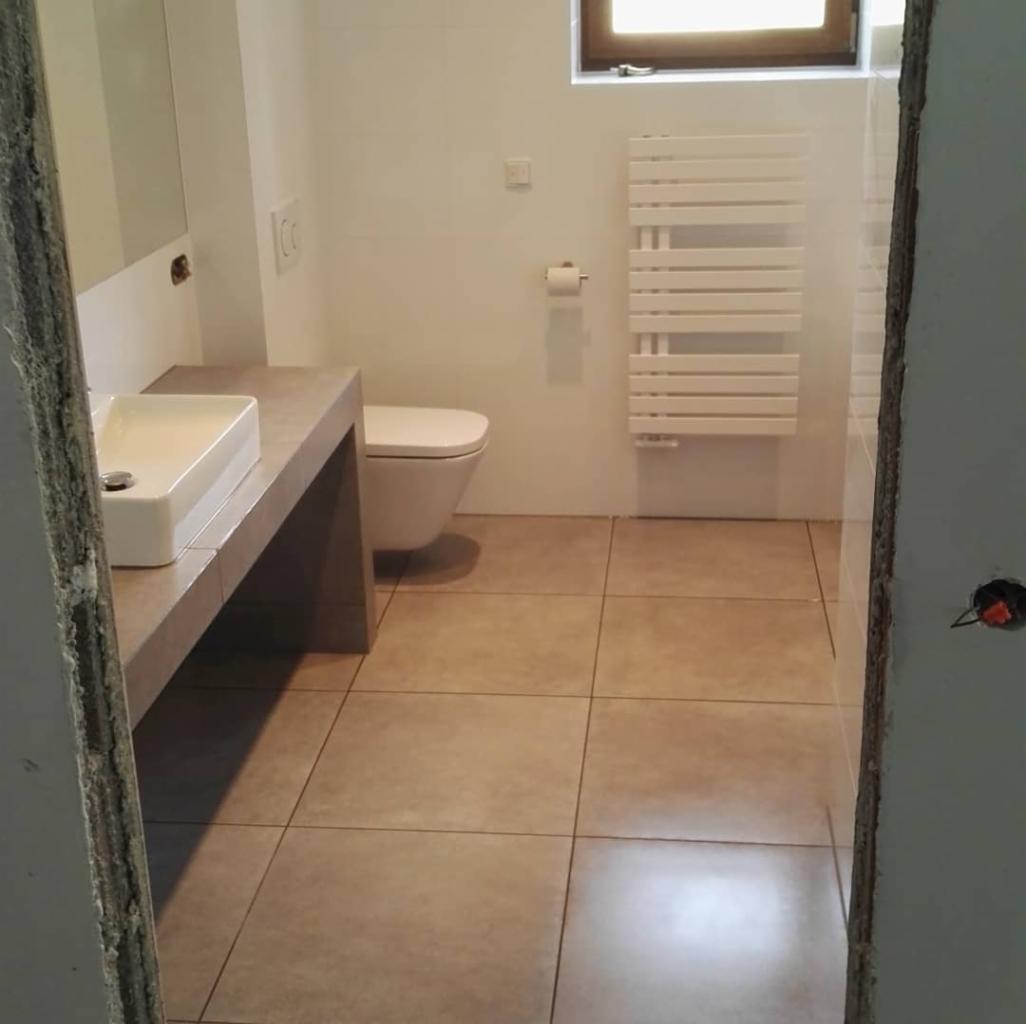 Ukończona łazienka w neutralnych kolorach: beżowe płytki podłogowe, biała umywalka na betonowym blacie, toaleta podwieszana i grzejnik drabinkowy na białej ścianie, widoczne ślady po remoncie...