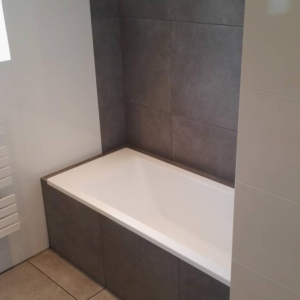 Nowoczesna łazienka z wanną obudowaną ciemnoszarymi płytkami, ściany wyłożone białą i szarą glazurą, widoczny fragment grzejnika drabinkowego.