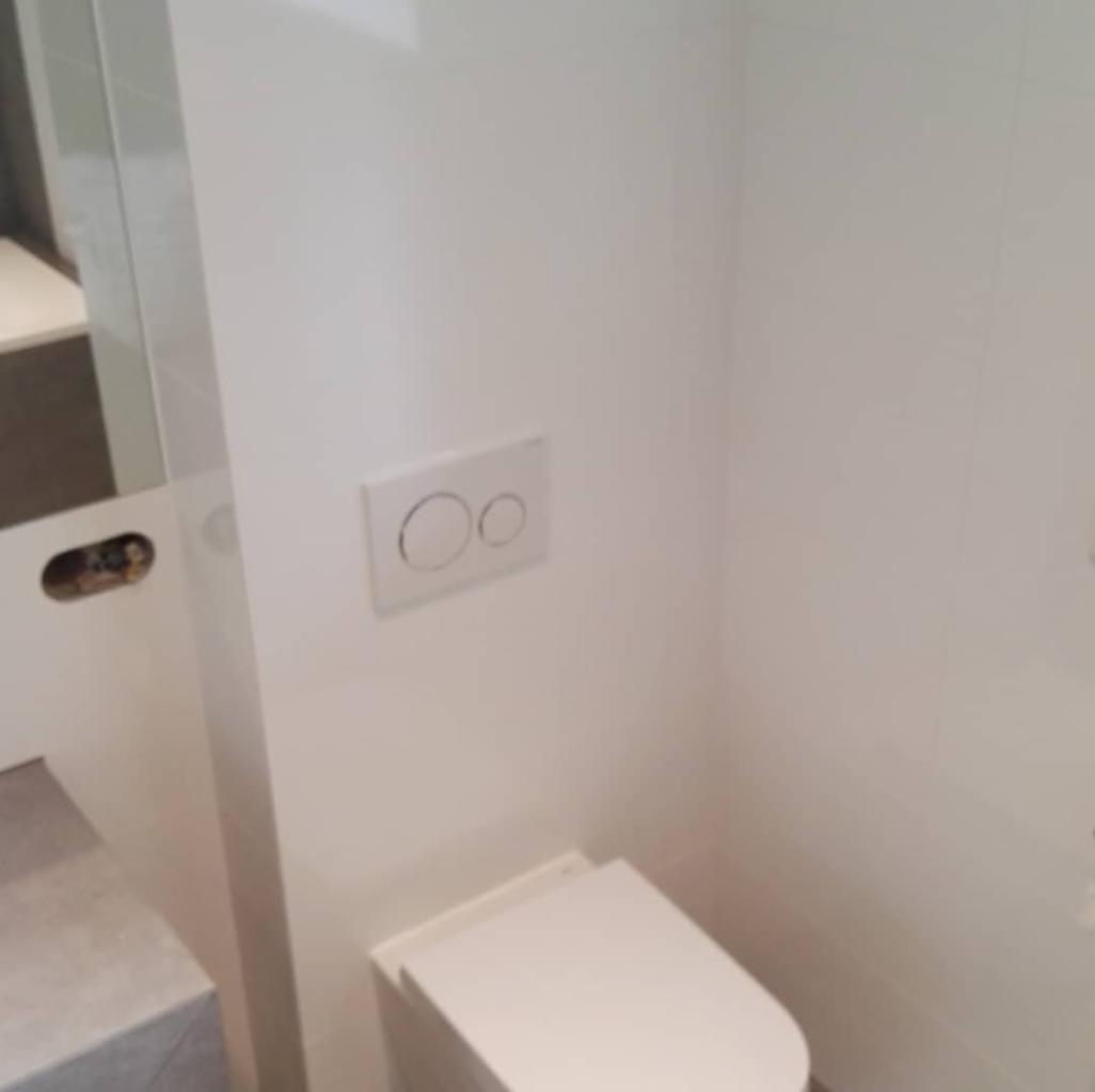 Biała toaleta podwieszana z przyciskiem spłukującym na ścianie wyłożonej białymi płytkami w wyremontowanej łazience.