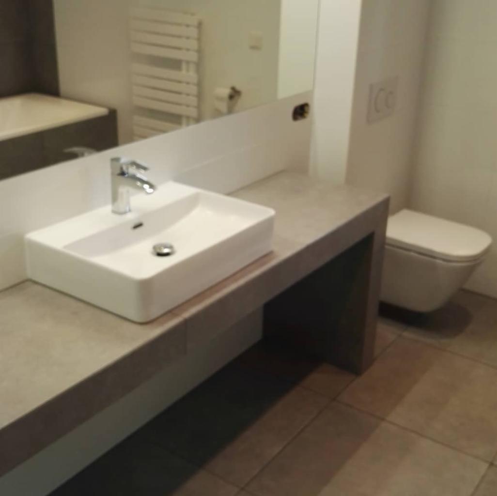 Nowoczesna łazienka z prostokątną umywalką nablatową, chromowaną baterią i toaletą podwieszaną, widok z boku na szaro-beżowe płytki podłogowe.