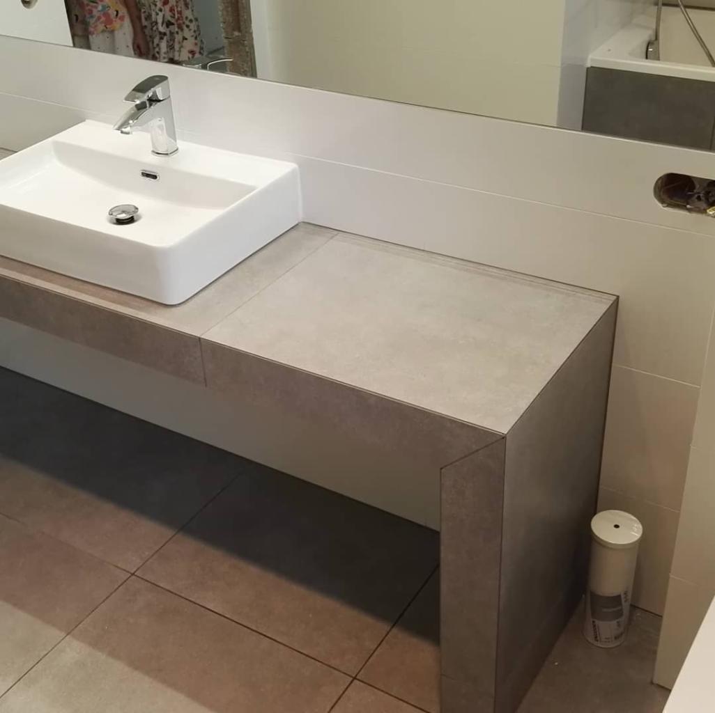 Nowoczesna łazienka: biała umywalka nablatowa z chromowaną baterią, szara zabudowa pod umywalką wykończona płytkami, szara podłoga z płytek imitujących kamień, widoczny fragment lustra i ściany...