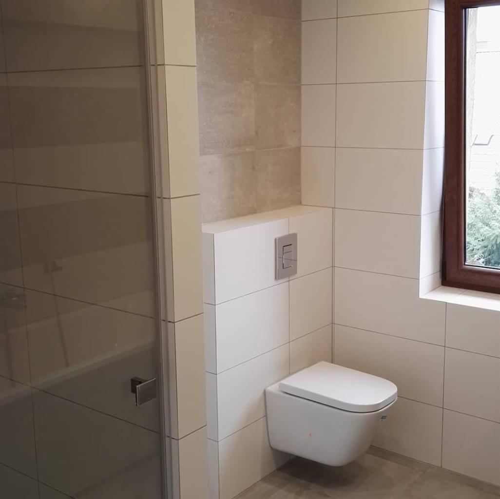 Nowoczesna łazienka z białymi, prostokątnymi płytkami, podwieszaną toaletą i szklaną kabiną prysznicową, widok na okno z brązową ramą i zielenią na zewnątrz.