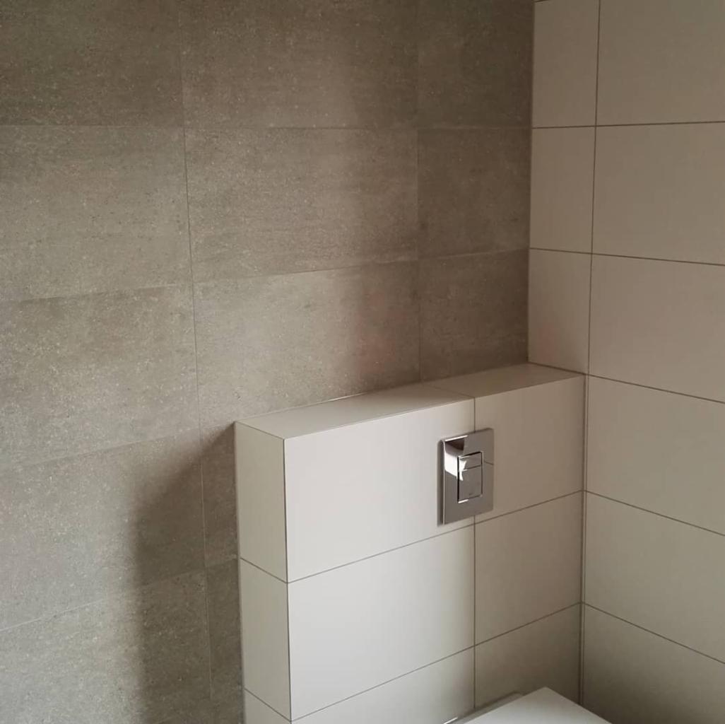 Wykończona ściana w łazience z szarymi płytkami imitującymi beton oraz biała zabudowa stelaża WC z chromowanym przyciskiem spłukującym.