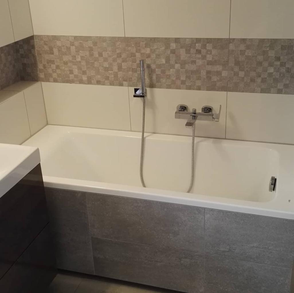 Wykończona łazienka w Rybniku: biała wanna obudowana szarymi płytkami imitującymi kamień, bateria wannowa chromowana, mozaika na ścianie w odcieniach beżu i szarości.