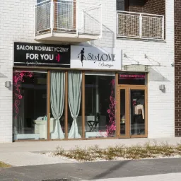 Fasada budynku z dwoma salonami kosmetycznymi, jeden z dekoracyjnymi naklejkami kwiatowymi na szybach, drugi z żółto-zielonym szyldem apteki obok.