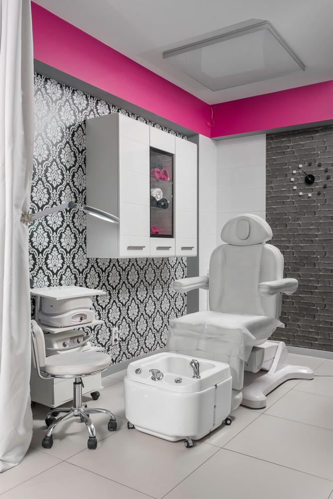 Nowoczesny gabinet kosmetyczny z białym fotelem do zabiegów, urządzeniem do pielęgnacji, wanną do pedicure na kółkach i szafką z akcesoriami, ściana w czarno-białe wzory i zegar z cegieł.