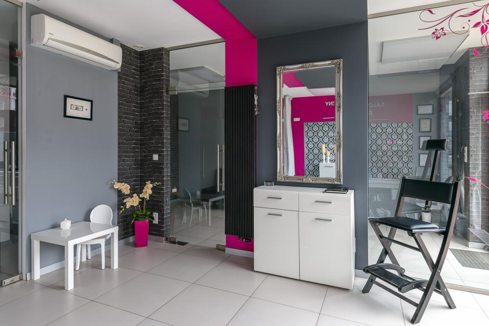 Nowoczesne wnętrze salonu: szare ściany z akcentem fuksji, białe meble, designerskie krzesło do wizażu i lustro w ozdobnej ramie.