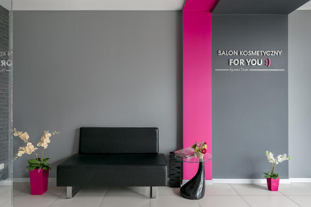 Wnętrze salonu kosmetycznego z czarną kanapą, szarą ścianą z różowym akcentem i napisem 'Salon Kosmetyczny For You', kwiatami w różowych doniczkach i stolikiem ze szklanym blatem.