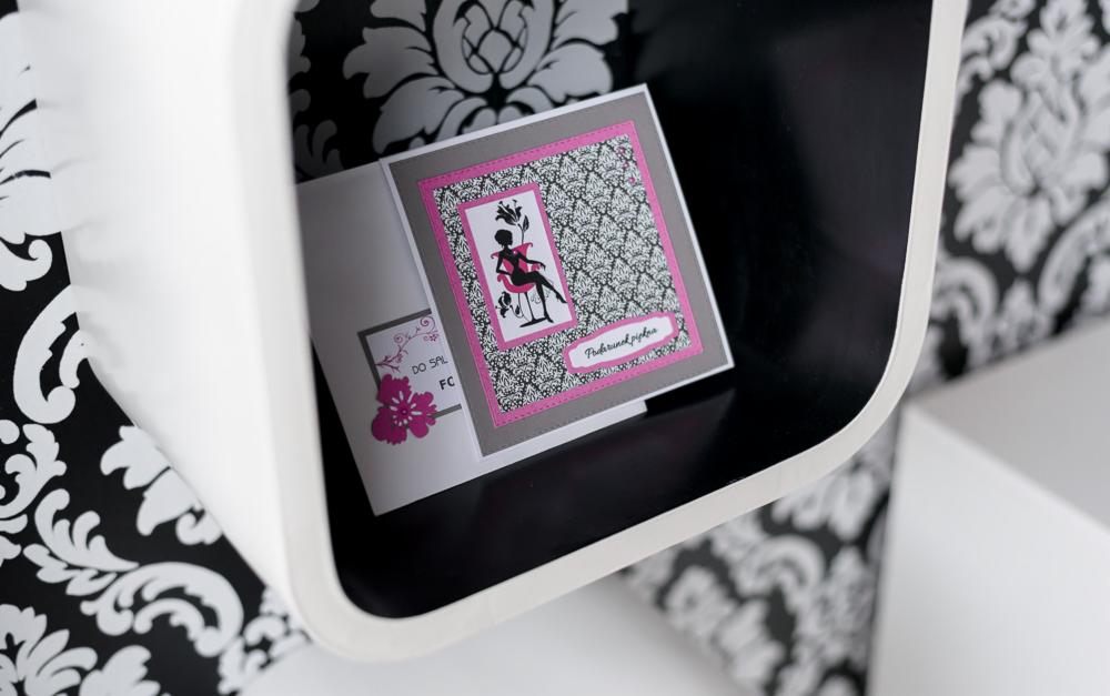 Ręcznie robiona kartka okolicznościowa z motywem kobiety, umieszczona w białej, zaokrąglonej półce na tle tapety w kwiatowe wzory.