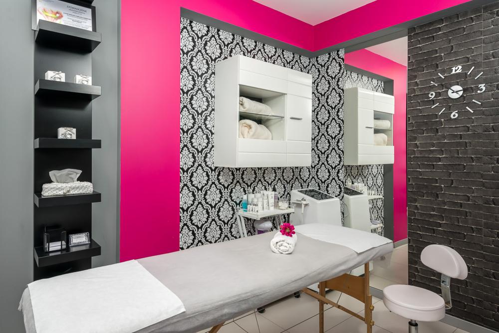 Wnętrze gabinetu kosmetycznego z łóżkiem do zabiegów, szafkami na kosmetyki i dekoracyjną ścianą w cegły z zegarem.
