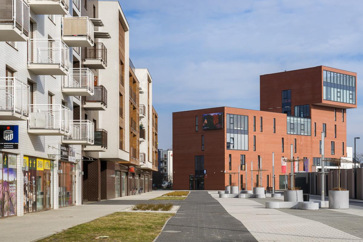 Nowoczesny kompleks budynków z białymi balkonami i ceglaną elewacją, widok z poziomu chodnika z elementami zieleni i betonowymi siedziskami.