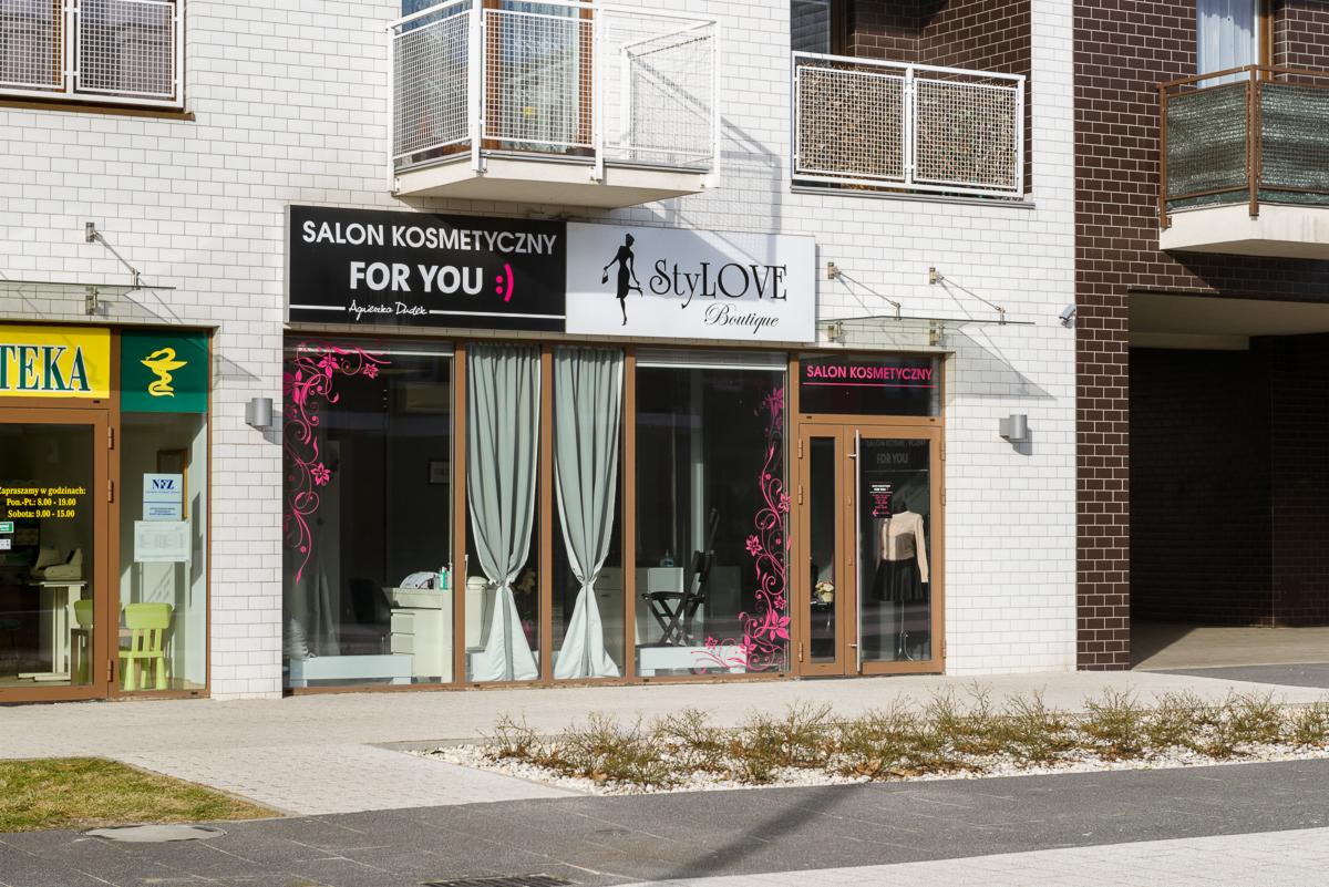 Fasada budynku z dwoma salonami kosmetycznymi, jeden z dekoracyjnymi naklejkami kwiatowymi na szybach, drugi z żółto-zielonym szyldem apteki obok.