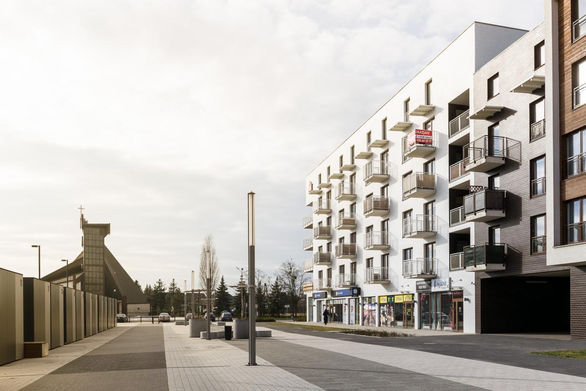Nowoczesny budynek apartamentowy z balkonami i lokalami usługowymi na parterze, widok z poziomu chodnika w słoneczny dzień, w tle kościół.