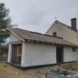 Wznoszenie ścian parterowego domu z bloczków, widoczne drewniane elementy konstrukcji dachu, czarna hydroizolacja fundamentów, w tle dom jednorodzinny.