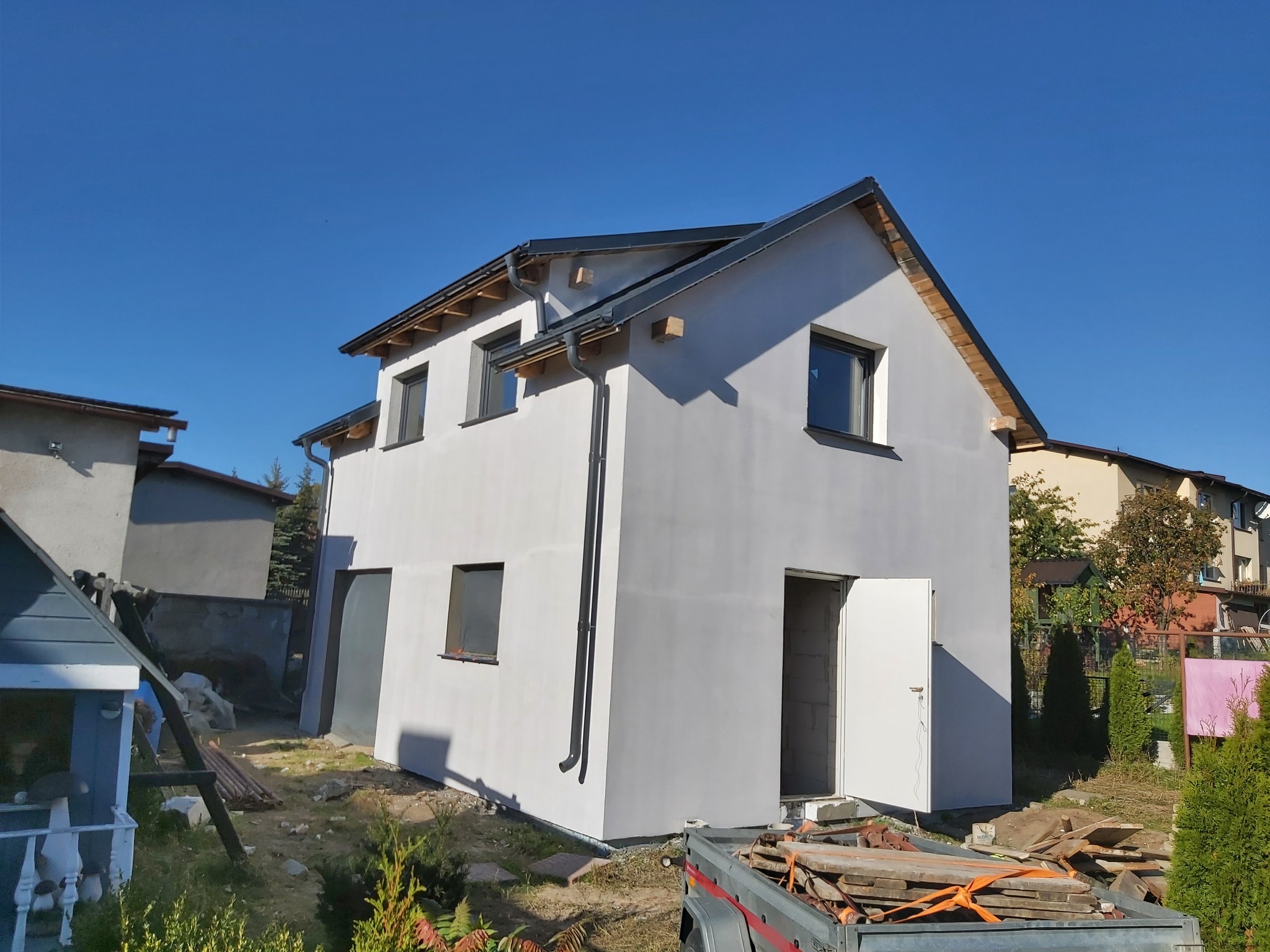 Dwukondygnacyjny, otynkowany dom w trakcie budowy, z widoczną przyczepką pełną materiałów budowlanych i otwartymi drzwiami wejściowymi, na tle błękitnego nieba.