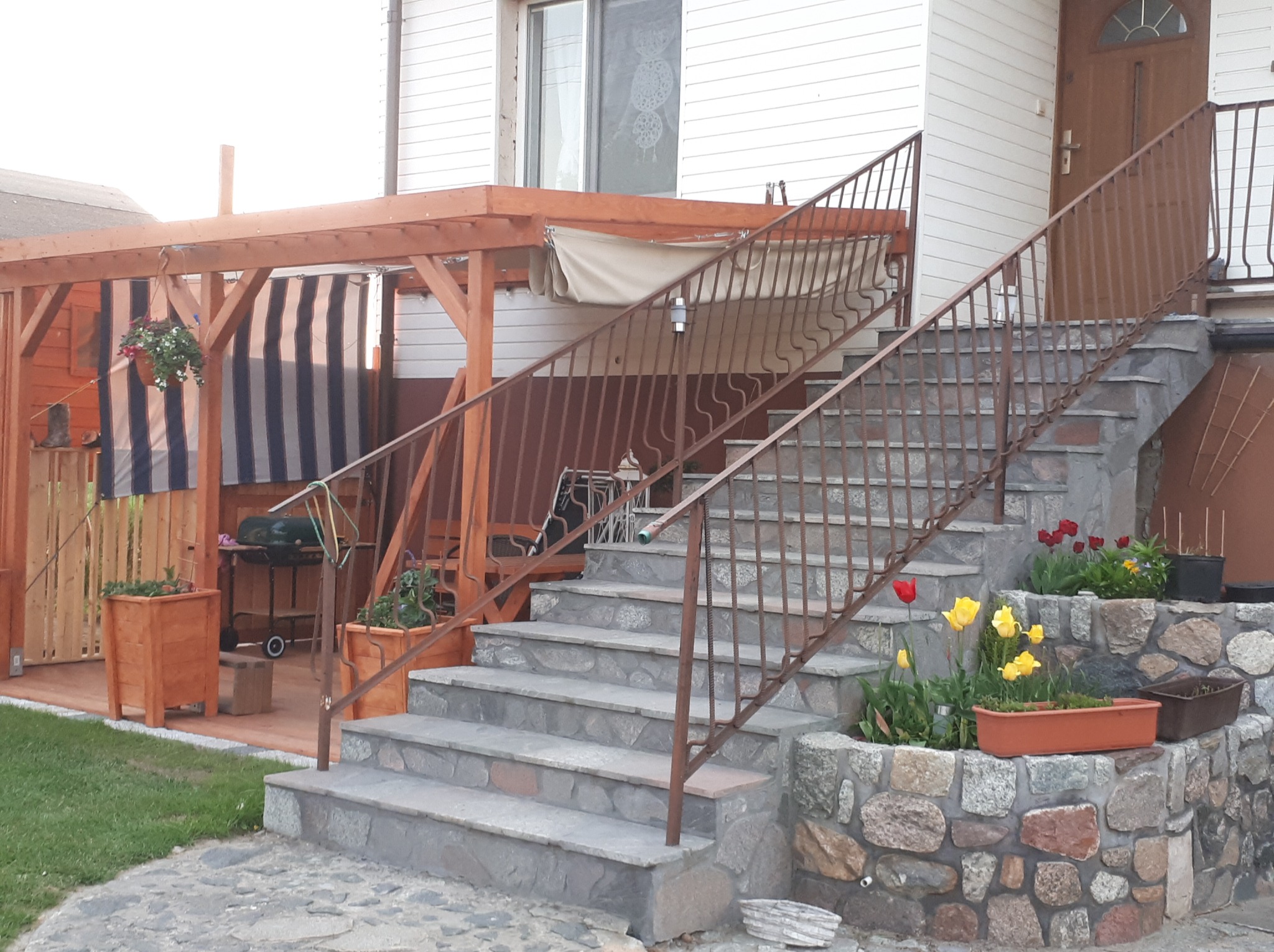 Schody wejściowe wyłożone kamieniem prowadzące do domu z drewnianą pergolą i grillem w tle.