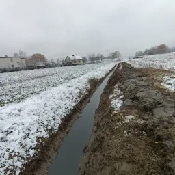 Wykonany drenaż liniowy na zewnątrz, w zasnieżonym terenie, widoczne domy w tle, pochmurne niebo.