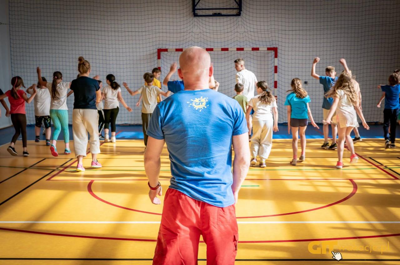 Grupa dzieci i dorosłych w sali gimnastycznej, prowadzący w czerwonych spodenkach i niebieskiej koszulce z nadrukiem na plecach, ćwiczenia grupowe z podniesionymi rękami, siatka do piłki ręcznej...