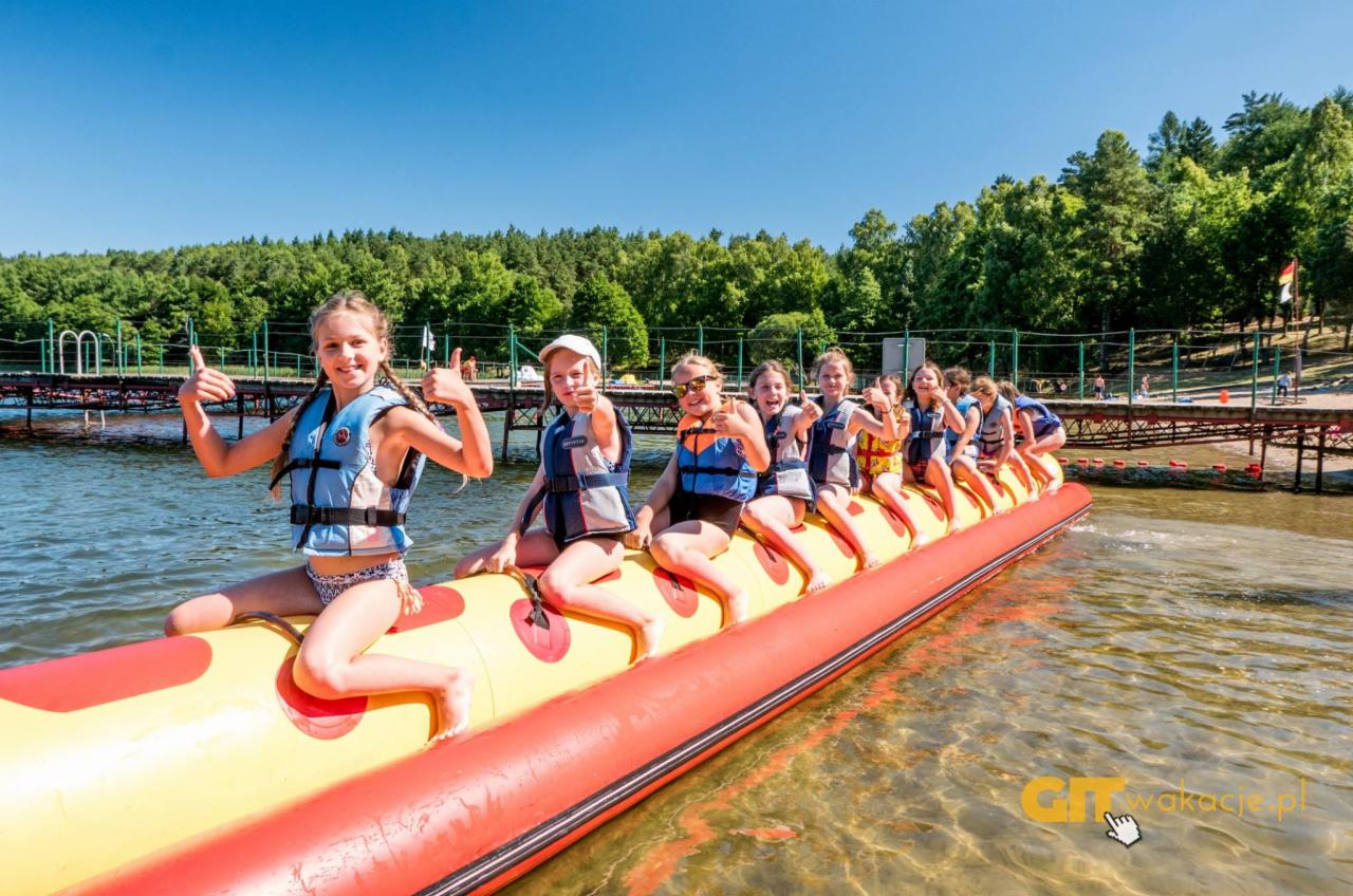 Grupa dzieci w kamizelkach ratunkowych siedzi na dmuchanym bananie na wodzie, z lasem w tle i pomostem w oddali.