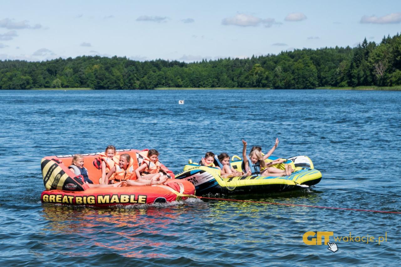 Grupa dzieci w kamizelkach ratunkowych na dmuchanych pontonach ciągniętych po jeziorze motorówką, w tle zielony las.