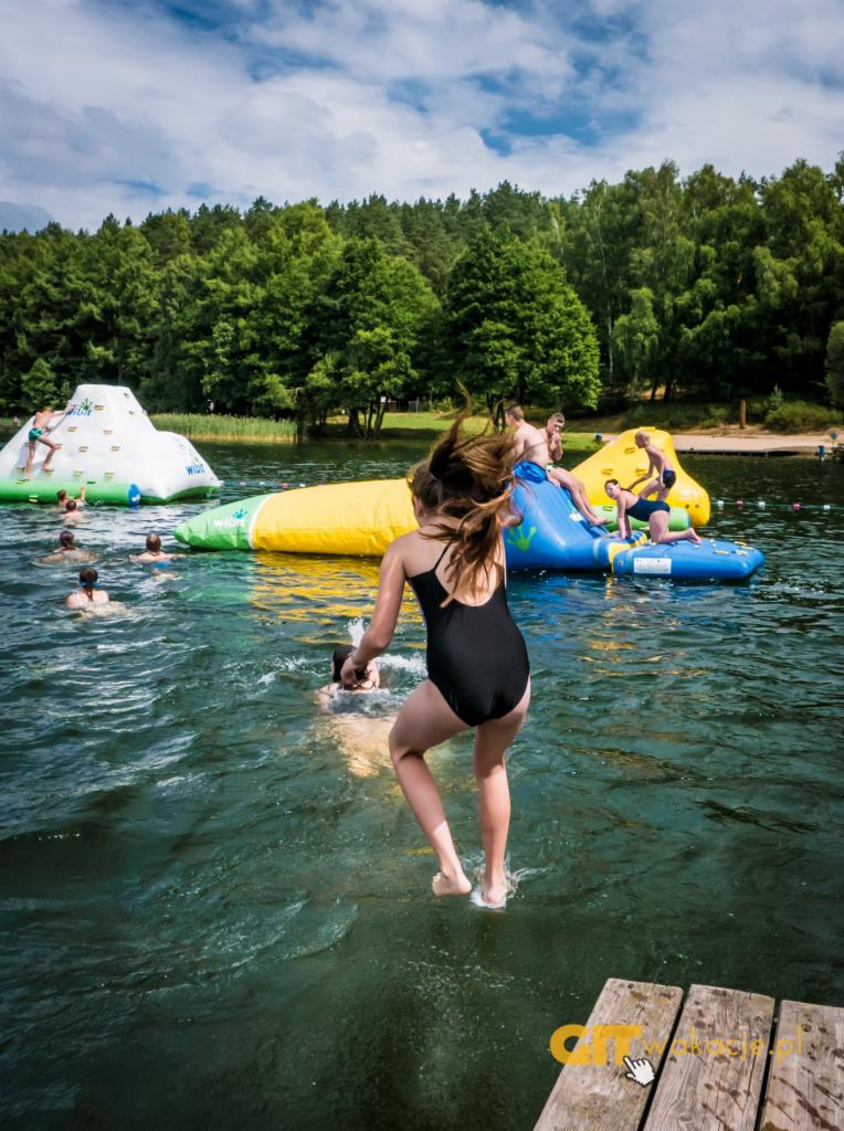 Dziewczynka w czarnym stroju kąpielowym skacze do jeziora z drewnianego pomostu, w tle dmuchane konstrukcje do zabawy i las.