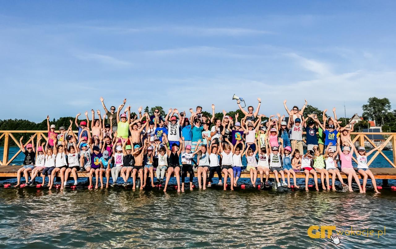 Duża grupa dzieci i opiekunów siedzi na drewnianym pomoście nad jeziorem, wszyscy z podniesionymi rękami, w tle błękitne niebo i zieleń drzew.