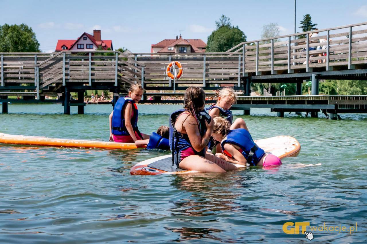 Grupa dzieci w kamizelkach ratunkowych siedzi na deskach SUP na jeziorze podczas kolonii letnich, w tle drewniany pomost i budynki.