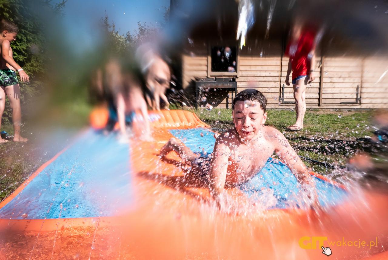 Chłopiec z zamkniętymi oczami zjeżdża na dmuchanej zjeżdżalni wodnej w Gdańsku, w tle widoczni inni uczestnicy zabawy i drewniany budynek.