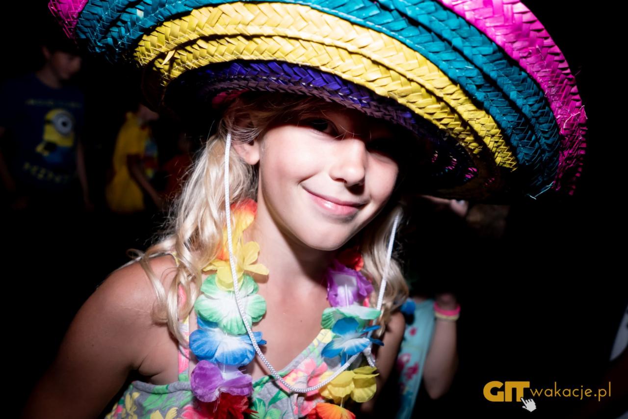 Uśmiechnięta dziewczynka w kolorowym sombrero i hawajskim naszyjniku z kwiatów na tle ciemnego otoczenia, prawdopodobnie podczas tematycznej imprezy.