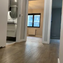 Widok z korytarza na wykończoną łazienkę z owalnym lustrem i salon z grzejnikiem pod oknem, podłoga wyłożona panelami imitującymi drewno.