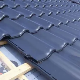 Układanie błyszczącej, grafitowej dachówki ceramicznej na łatach, widoczny fragment komina w tle.