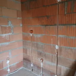 Instalacja elektryczna podtynkowa w trakcie montażu w surowym wnętrzu z czerwonej cegły dziurawki, widoczne kable w peszlach i puszki instalacyjne.