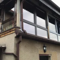 Zewnętrzny widok przeszklonej werandy z drewnianymi ramami okiennymi, z widocznymi śladami pianki montażowej i prac remontowych, na tle fragmentu elewacji budynku i sąsiedniego dachu.