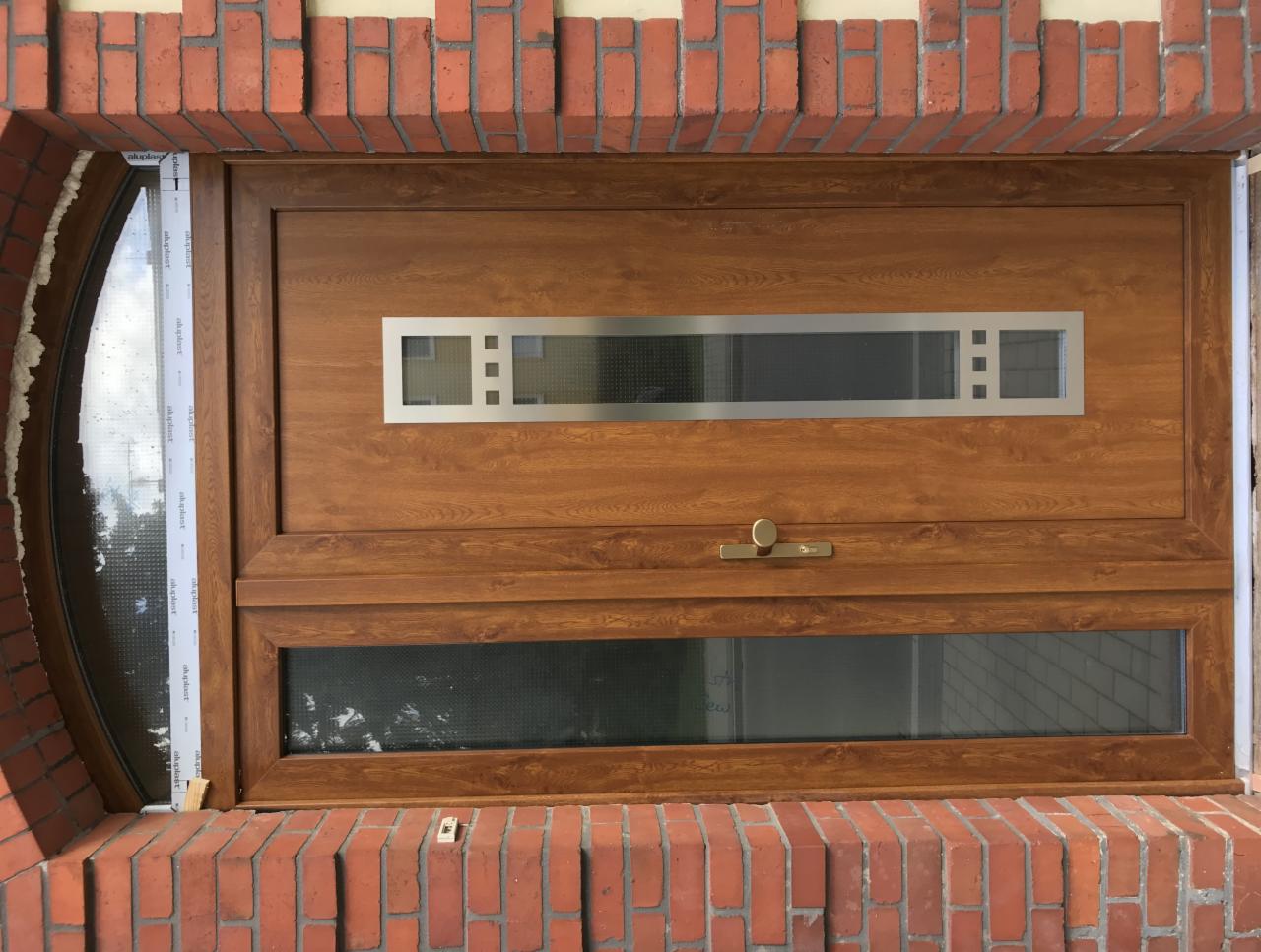 Brązowe okno z dwoma poziomymi przeszkleniami i okrągłym oknem bocznym, osadzone w czerwonej ceglanej elewacji. Widoczne ślady pianki montażowej.