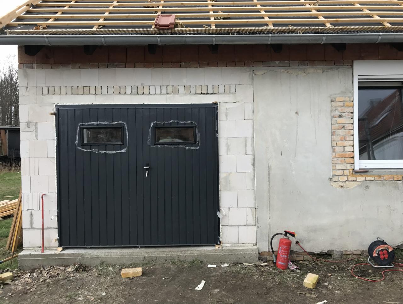 Czarna brama garażowa z dwoma małymi okienkami, zamontowana w surowym murze z betonu komórkowego, z widocznym oknem po prawej stronie i elementami konstrukcji dachu powyżej.