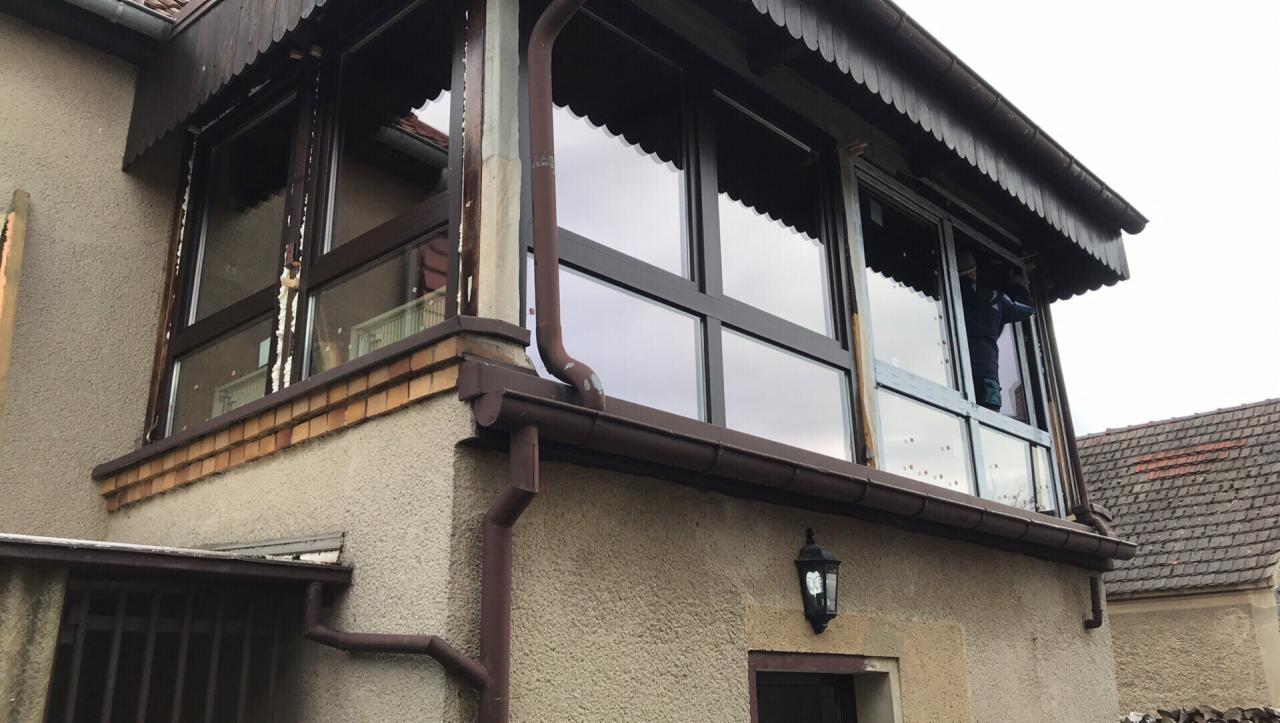 Zewnętrzny widok przeszklonej werandy z drewnianymi ramami okiennymi, z widocznymi śladami pianki montażowej i prac remontowych, na tle fragmentu elewacji budynku i sąsiedniego dachu.