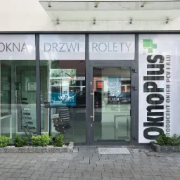 Fasada budynku firmy OknoPlus z dużymi przeszkleniami, na których widnieją napisy OKNA, DRZWI, ROLETY oraz logo firmy, przed wejściem zadbana zieleń w kamiennych donicach, widoczna kamera monitoringu.