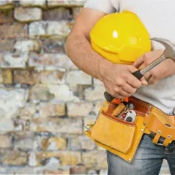 Murarz trzymający żółty kask i młotek, z pasem narzędziowym na tle ściany z cegły