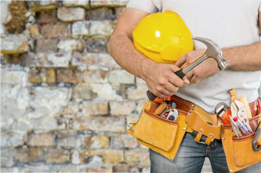 Murarz trzymający żółty kask i młotek, z pasem narzędziowym na tle ściany z cegły