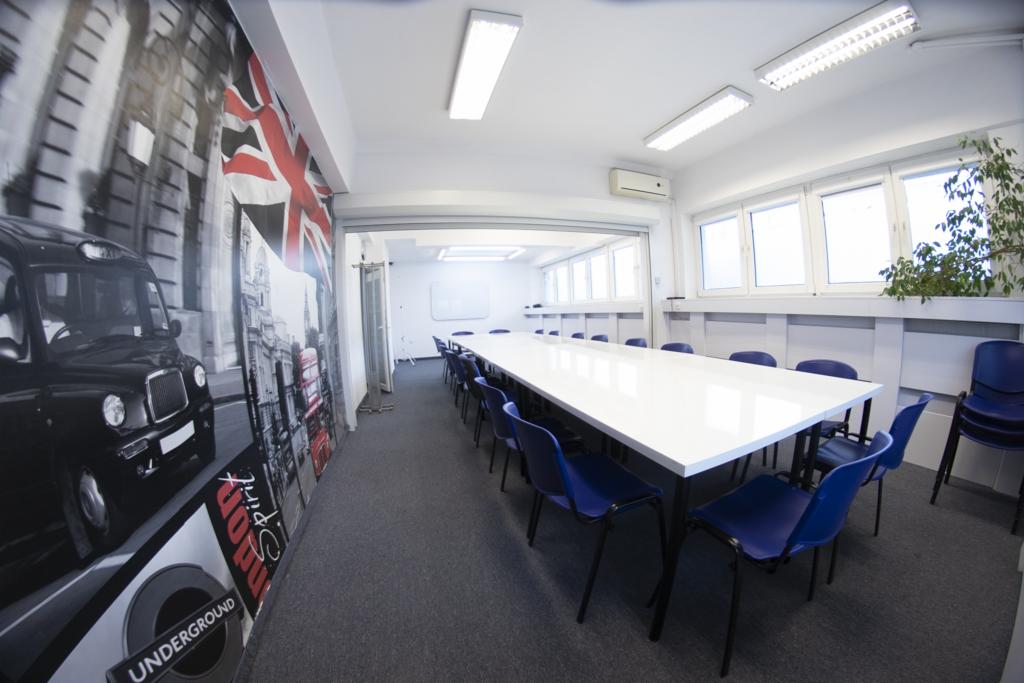 Sala szkoleniowa z długim białym stołem i niebieskimi krzesłami, na ścianie fototapeta z motywami londyńskimi: czarna taksówka, czerwony autobus i flaga Wielkiej Brytanii.